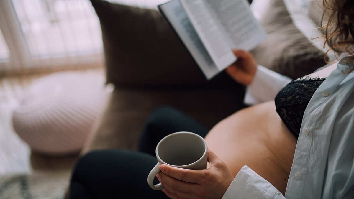 Femme enceinte assise sur un canapé, une tasse à la main, lisant un livre
