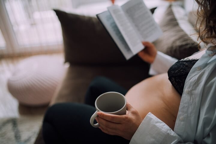 Femme enceinte assise sur un canapé, une tasse à la main, lisant un livre